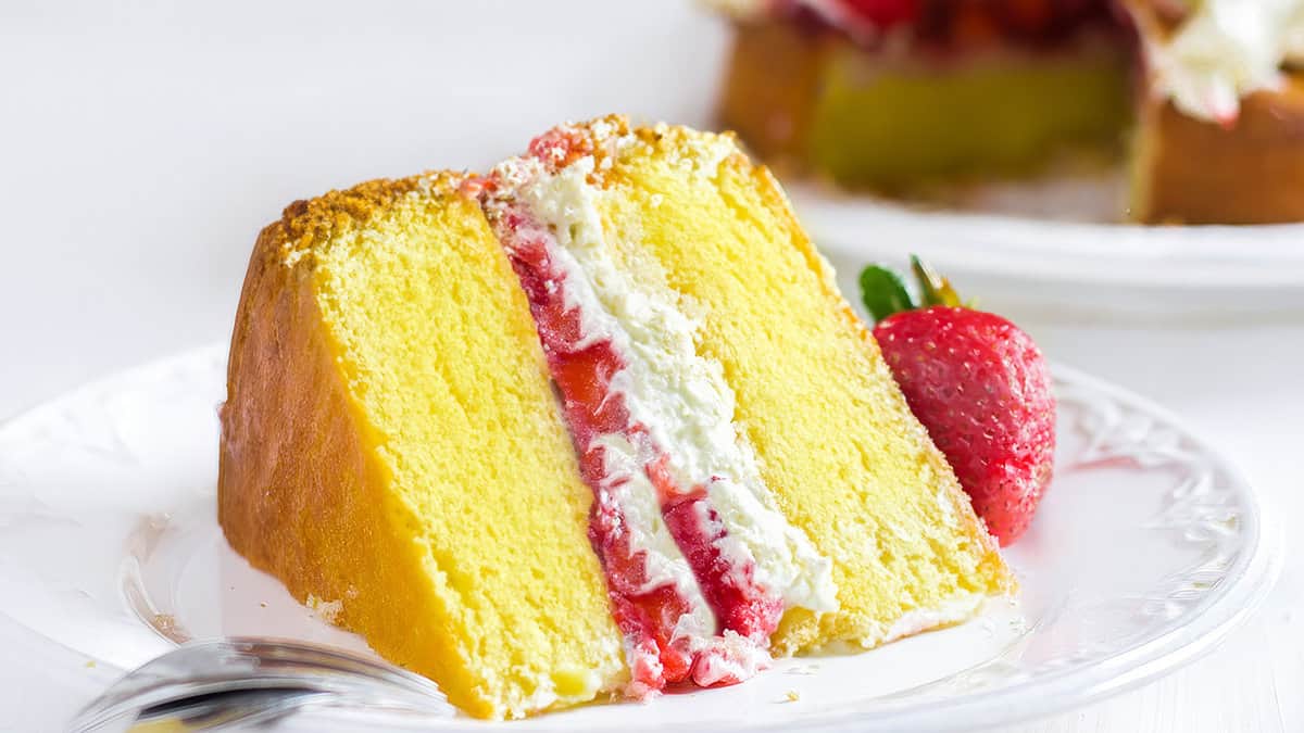 A slice of lemon cake filled with whipped cream and strawberries on a plate with the cake behind it.