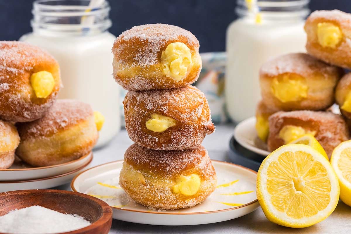 A stack of three sugar-coated bomboloni filled with yellow custard, surrounded by more custard-filled donuts, a halved lemon, a bowl of sugar, and milk jars with straws in the background.