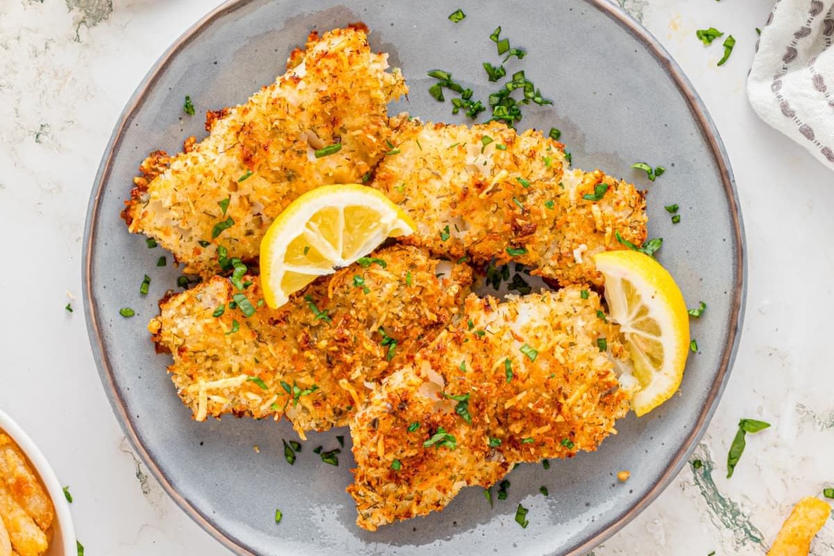 Breaded fish fillets from air fryer recipes garnished with lemon slices and chopped parsley on a gray plate.