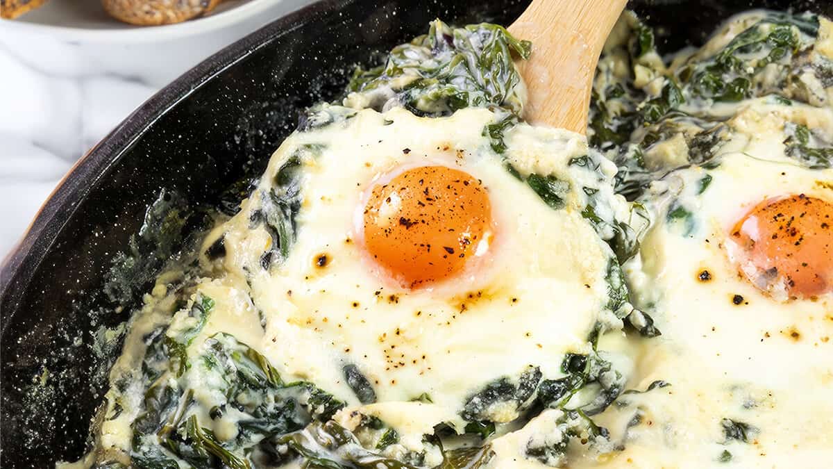 The photo shows baked Florentine eggs up close. It shows eggs baked in a skillet, with the yolk of an egg front and center, glossy and slightly runny. The eggs are topped with a rich mixture of melted cheese and creamy spinach.