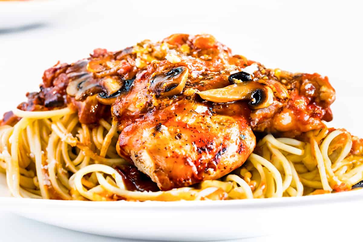 A close up image of Chicken Cacciatore over spaghetti with a glass of red wine in the background.