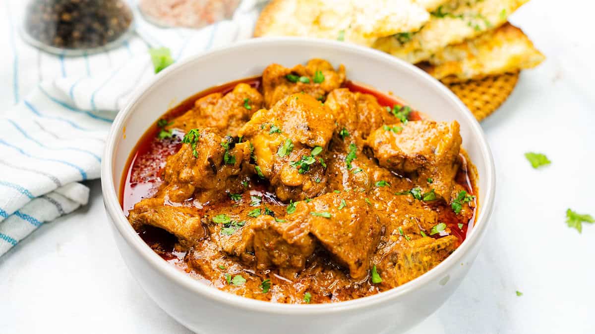 A heaping bowl of chicken korma, garnished with fresh coriander, sits next to garlic naan bread on a woven mat.