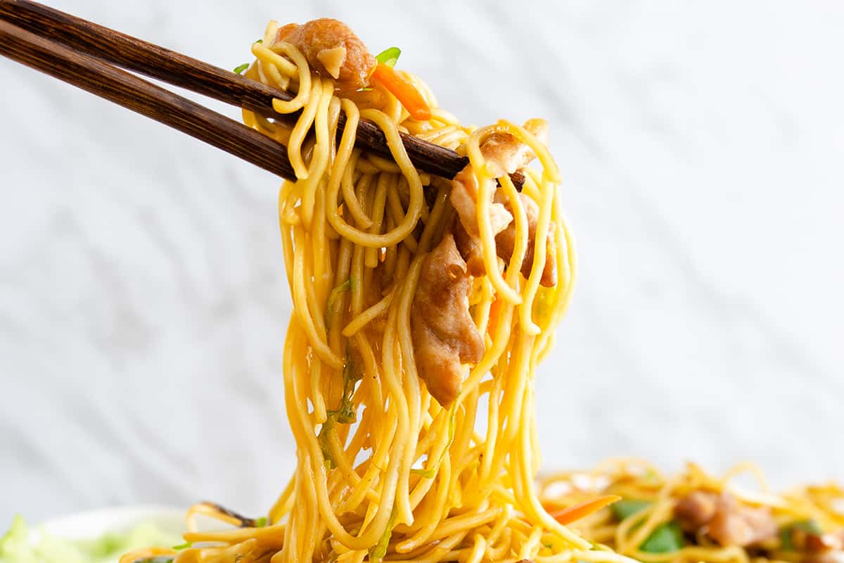 Chopsticks holding a strip of chicken lo mein with noodles, vegetables and chicken, on a blurred background.