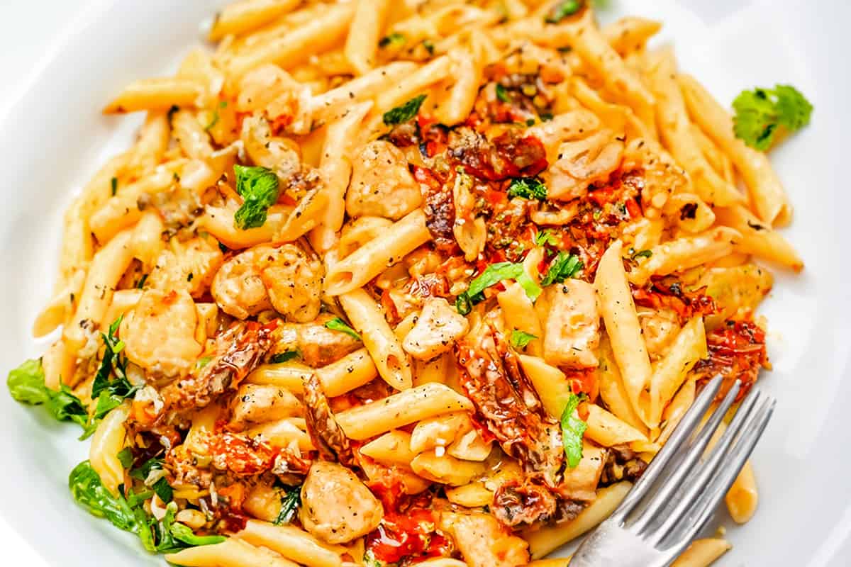 A white plate with chicken pasta with sun-dried tomatoes and a fork.