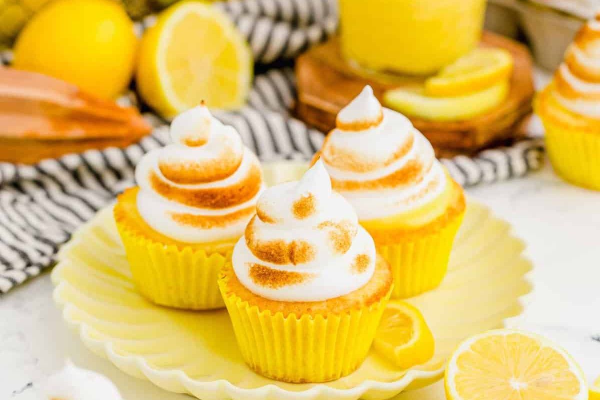 Three lemon cupcakes with toasted meringue frosting are arranged on a yellow plate, personifying delicious lemon desserts, with sliced ​​lemons and a striped cloth in the background.