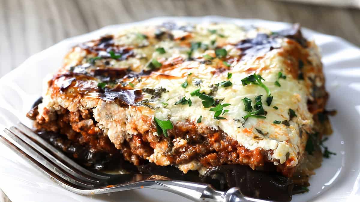 A slice of moussaka on a white plate and a fork next to it.