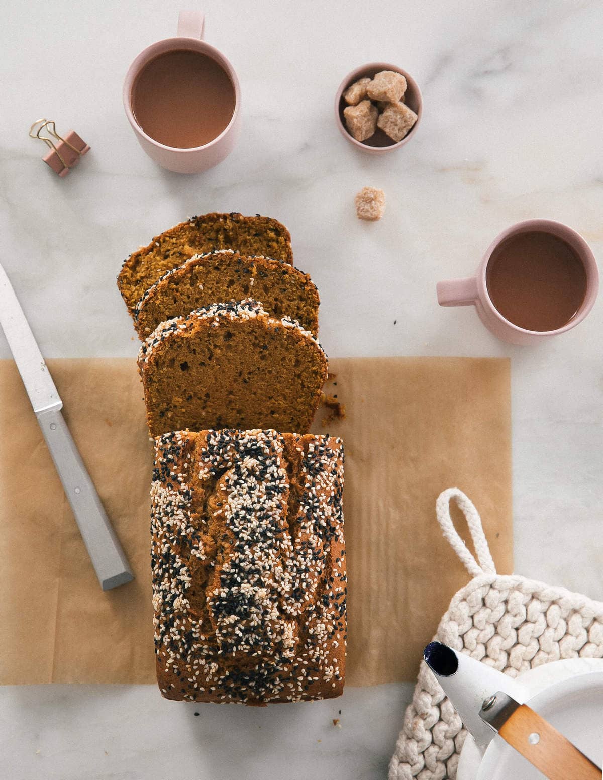 Overhead view of a pumpkin tahini bread on a counter with slices and coffee cups arranged around it.