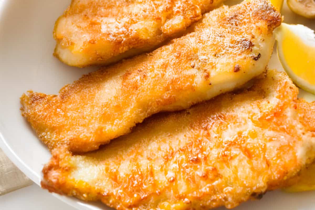 Pan-fried, golden fish fillets are neatly arranged on a white plate, displaying their crispy texture. A slice of lemon is placed next to them, adding a splash of color to the dish.