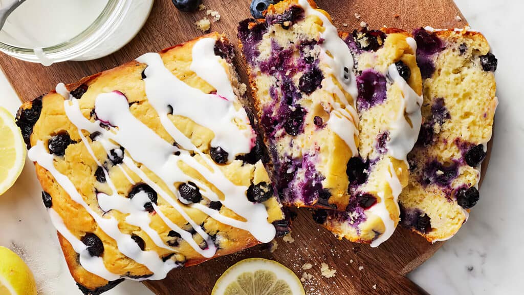 A loaf of blueberry lemon bread with white icing sits on a wooden cutting board. Two slices have been cut from the loaf, showing blueberries and a moist texture inside. A lemon slice and jar nearby add to the decorative presentation, perfect for exploring blueberry recipes.