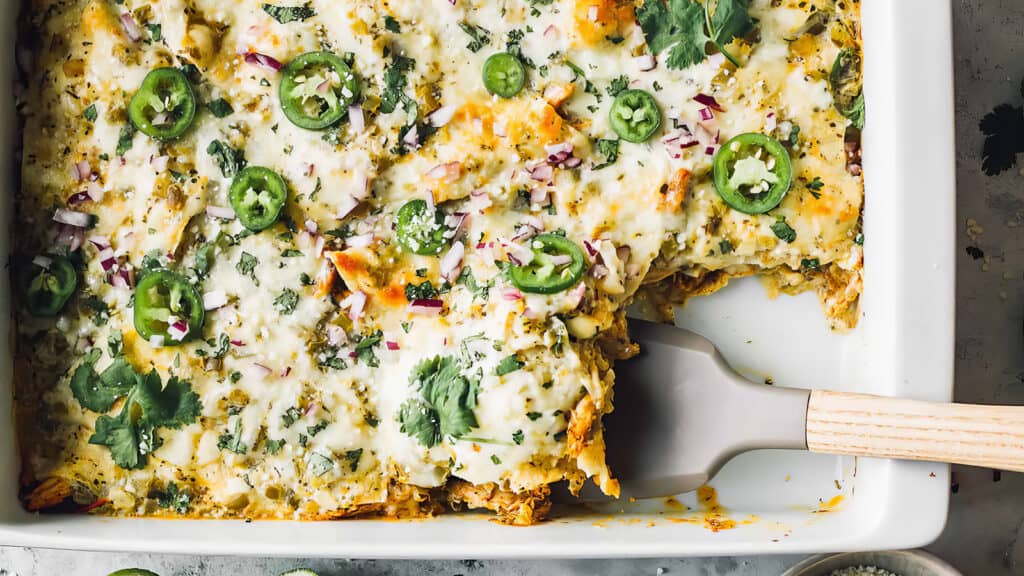 Shown is a rectangular white plate of delicious chicken casserole with green sauce, topped with melted cheese, sliced ​​jalapeños, chopped red onions, and cilantro. 