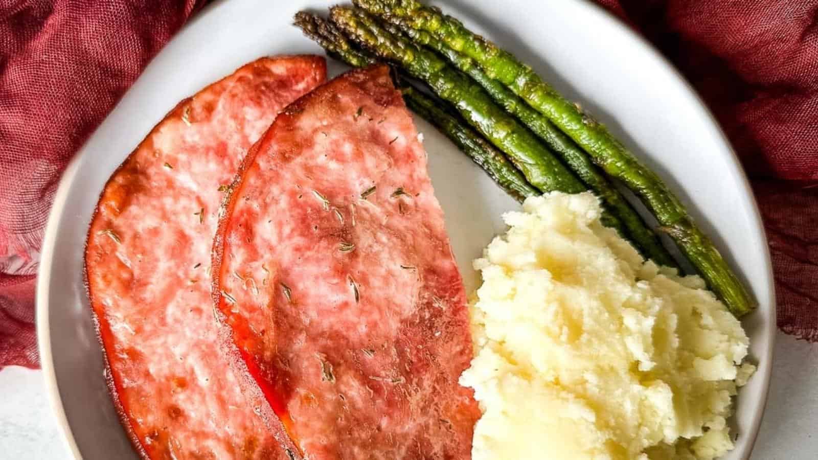 Air fryer ham steak on a white plate with asparagus and mashed potatoes.