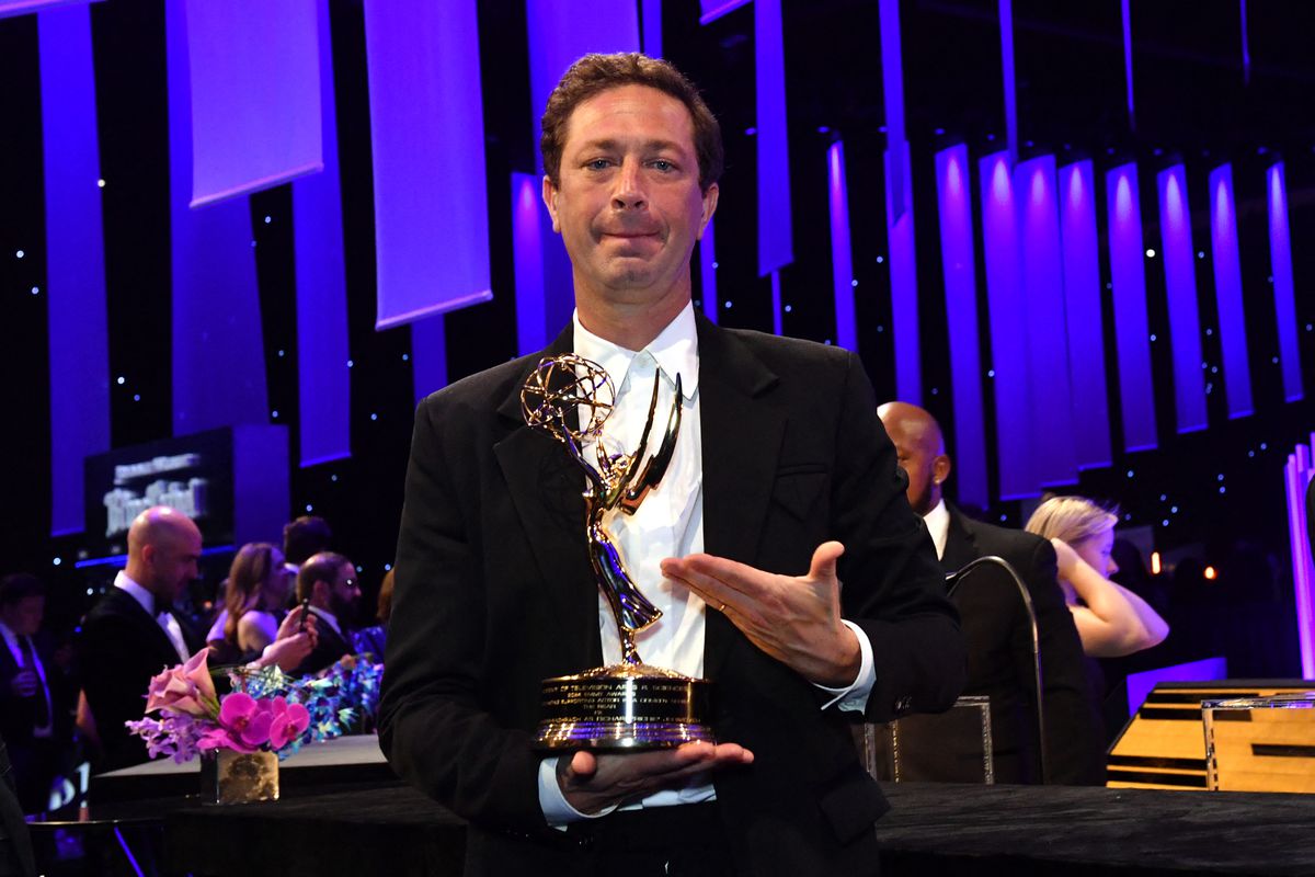 A man wearing a suit without a tie gestures toward an Emmy award in his hands inside an auditorium.