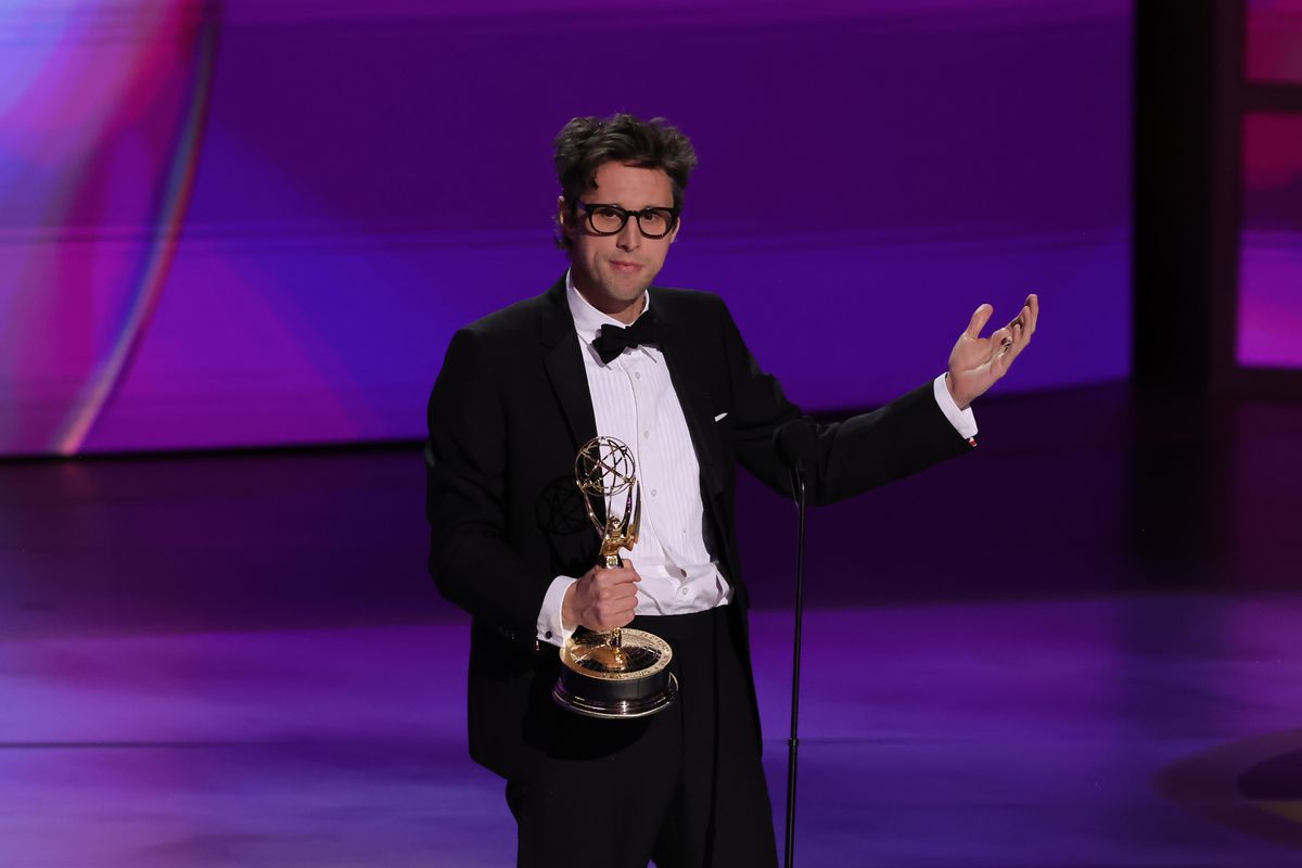 A man in glasses and a tuxedo on stage in front of a microphone, gesturing while holding an Emmy Award.