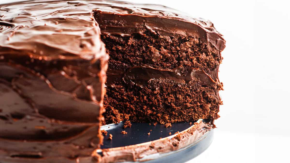 A devil's food cake with a slice removed, showing the rich cake layers and smooth chocolate frosting, on a white plate atop a dark slate board, against a marble background.