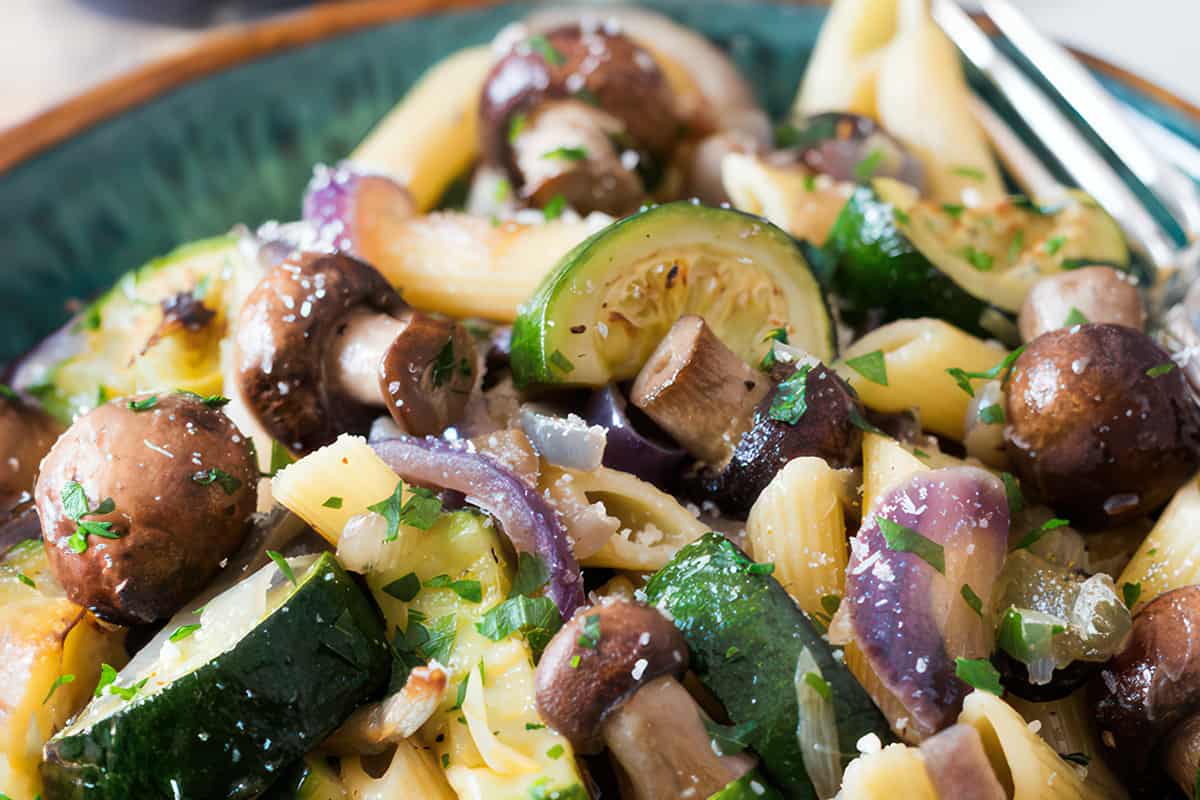 A close-up view of roasted vegetable pasta with zucchini, brown mushrooms, red onions and parsley over penne, sprinkled with coarse salt and grated cheese.