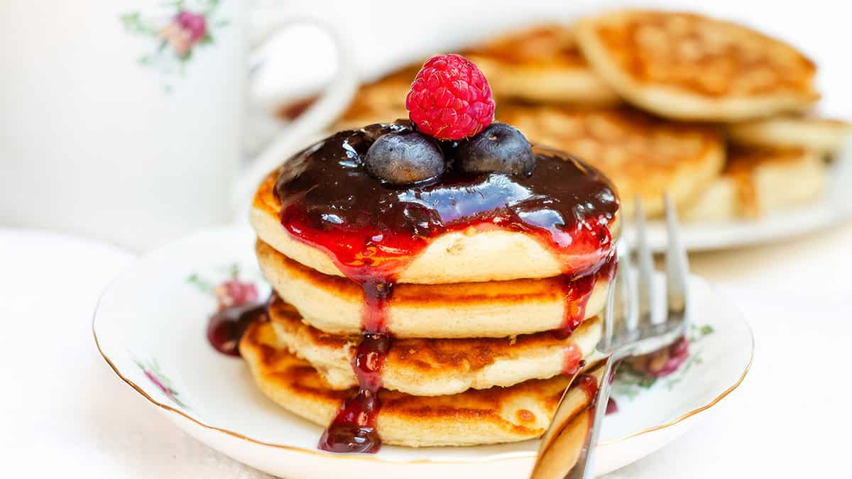 A stack of Scottish pancakes topped with a bright berry sauce and fresh berries, with a cup of tea and additional pancakes in the background, all on a white tablecloth.