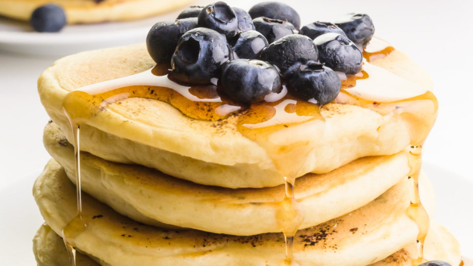 A stack of pancakes has blueberries on top and lots of maple syrup dripping down the sides.