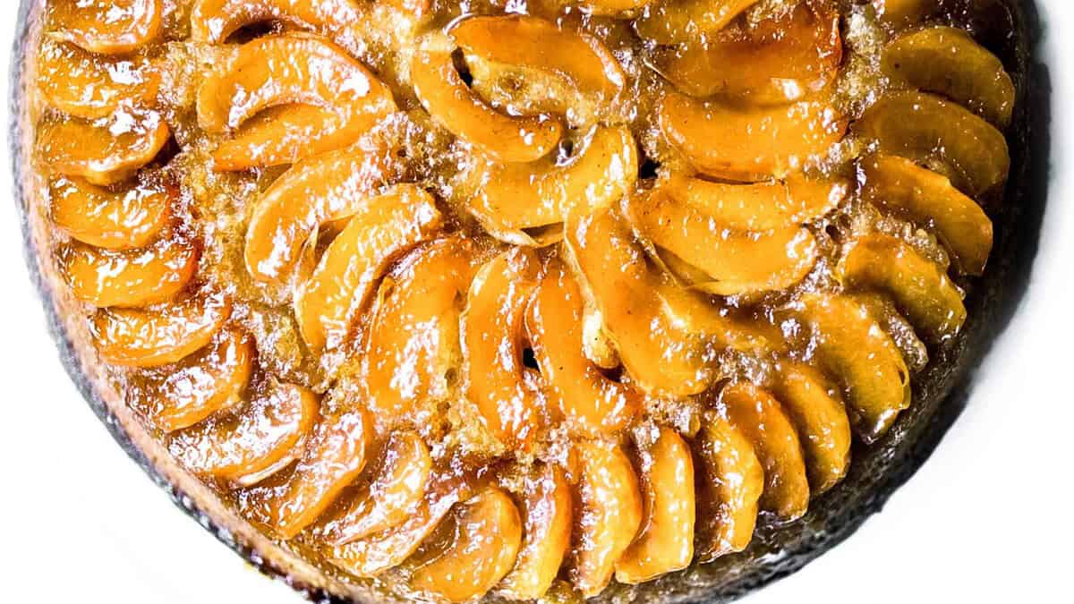 A golden upside-down cake with slices of caramelized apples neatly arranged on top. The glossy finish indicates a sugary glaze, making it an attractive and tempting dessert. Ideal for those exploring new dessert recipes, the cake appears moist and is probably freshly baked.