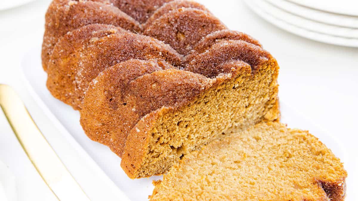 The image shows a loaf of bread with applesauce, golden brown and sugary crust, partially cut on a white plate next to a golden knife.