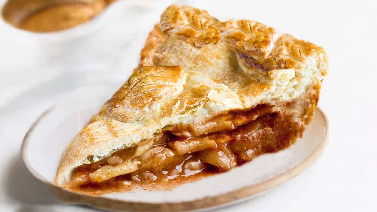 A closeup of a slice of apple pie on a white plate. The pie has a flaky, golden-brown crust with visible layers of baked apple slices and cinnamon, embodying the essence of Thanksgiving desserts. A small portion of sauce oozes out of the filling and the background is slightly blurry.