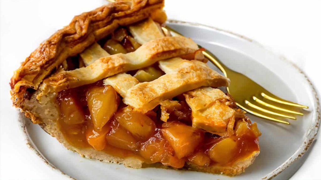 A close-up of a slice of peach cobbler, showing a golden brown lattice crust and a juicy peach filling, served on a white plate with a golden fork.