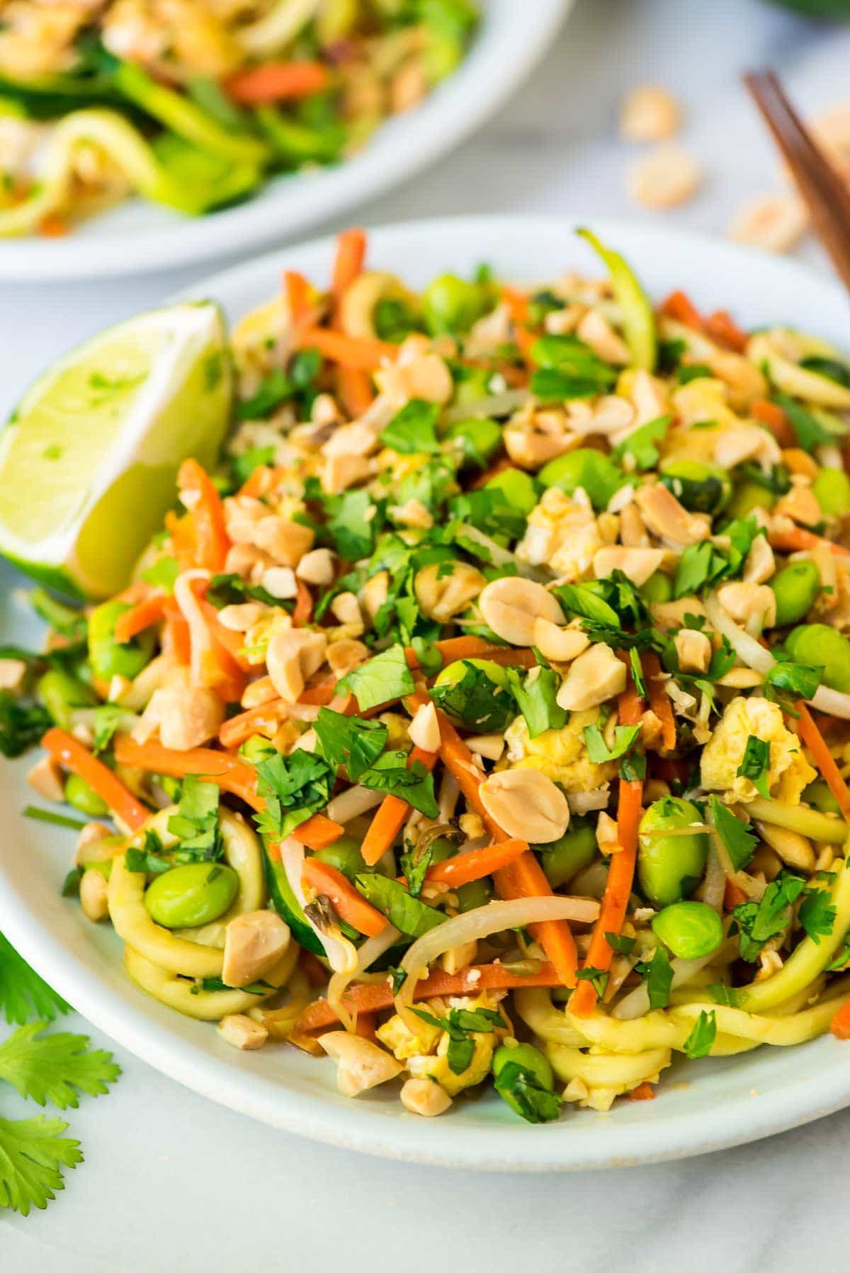 Zoodle Pad Thai in a white bowl with chopsticks and garnished with lime slices, cilantro and peanuts