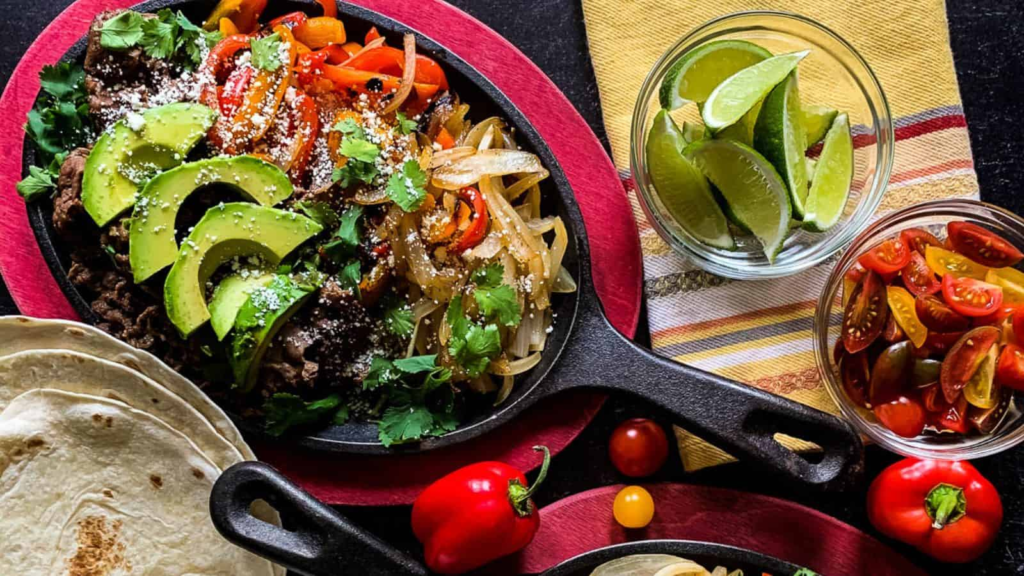 A cast iron skillet with hot fajita beef and seared vegetables along with bowls of lime wedges, tomatoes, and tortillas.